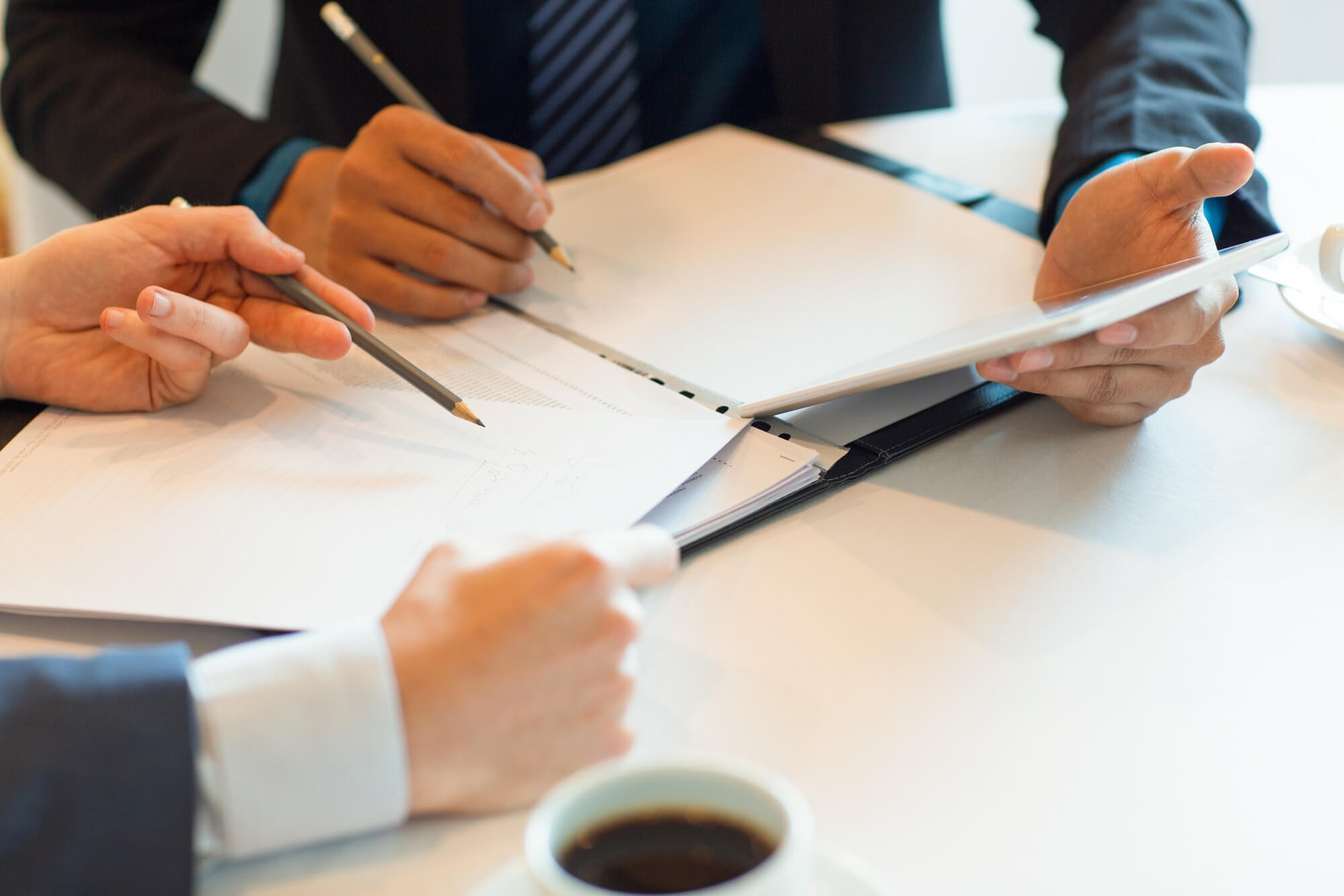 Three people in business attire review documents on a table with pencils and a tablet, alongside a cup of coffee.