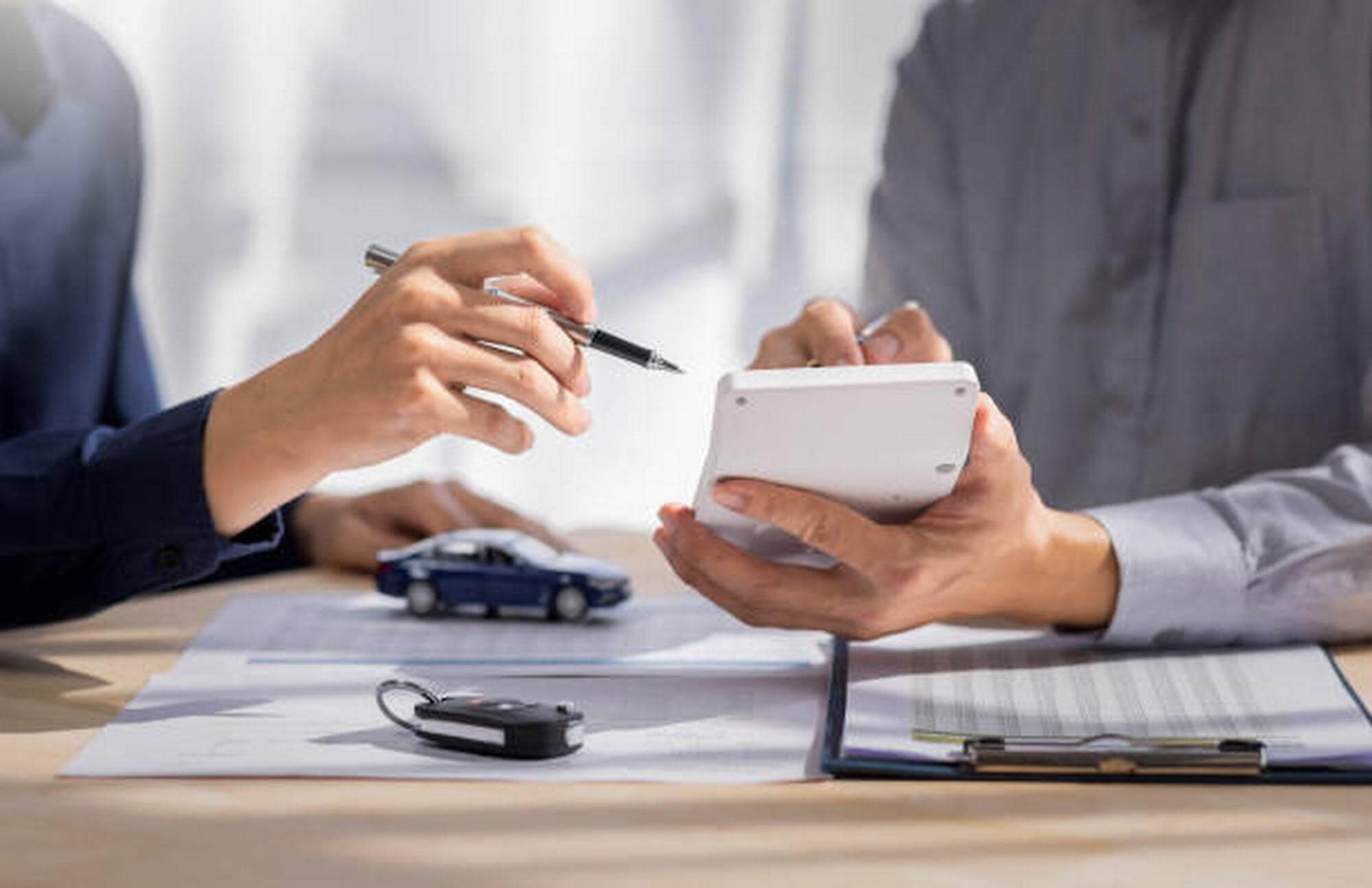 Two people discussing car insurance with a toy car, car key, and documents on the table while one holds a calculator.